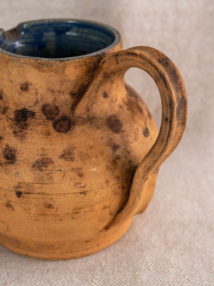 Antique French terracotta pitcher with curved handle, cobalt blue glazed interior, dark speckles, on beige fabric.