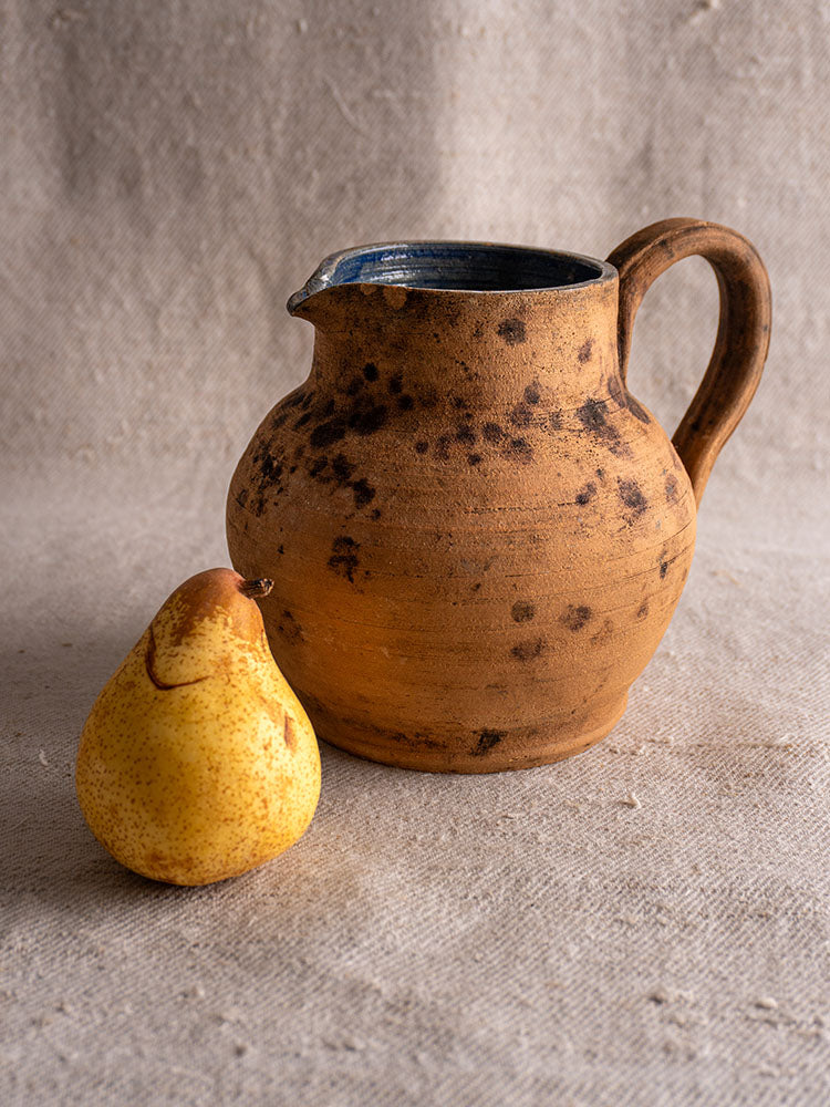 Antique French terracotta pitcher with cobalt blue glazed interior beside yellow pear on beige textured fabric.