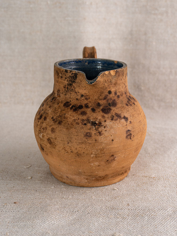 Antique French terracotta pitcher with wide body, cobalt blue glazed interior, on textured beige fabric.