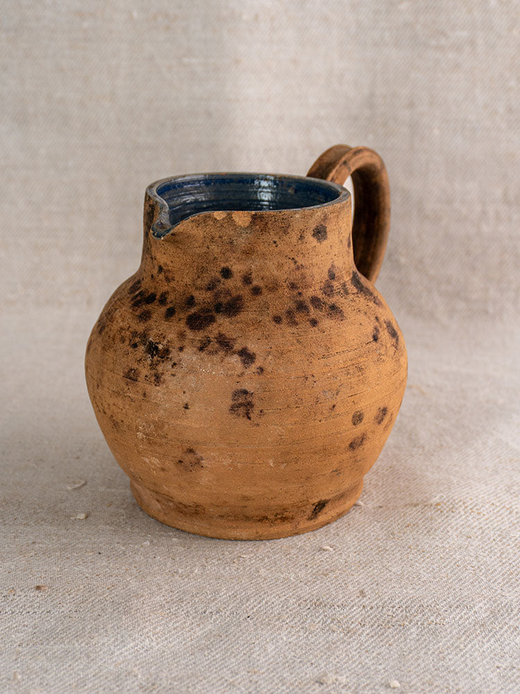 Mid-19th century French terracotta pitcher with rounded body, cobalt blue interior, brown ceramic glaze, on beige fabric.