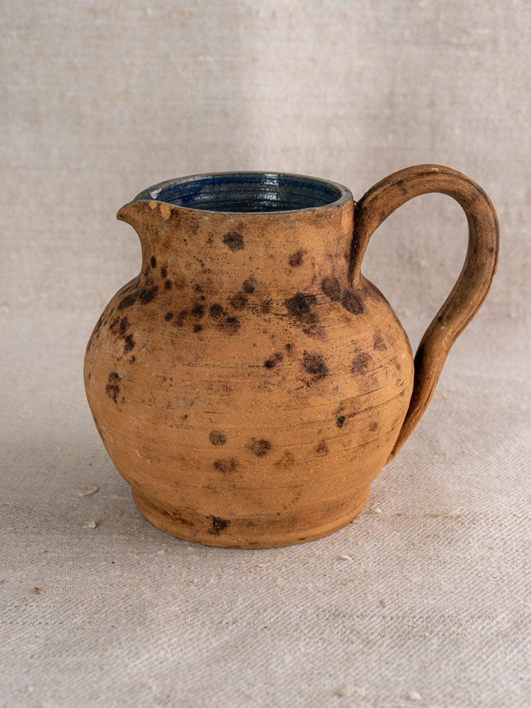 Antique French terracotta pitcher with wide body, curved handle, cobalt blue interior on textured beige fabric.