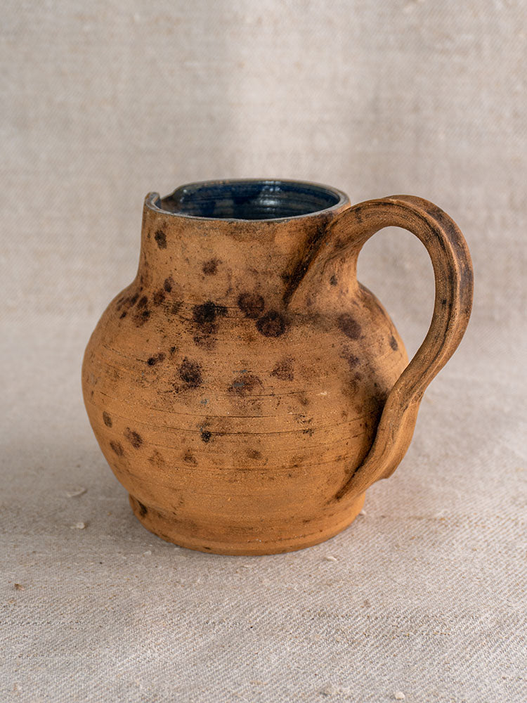 Antique French terracotta pitcher with cobalt blue glazed interior and curved handle, wide mouth, on textured fabric.