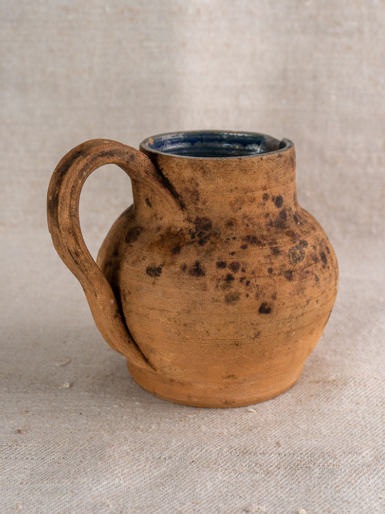 Antique French terracotta pitcher with curved handle and cobalt blue glazed interior on light fabric background.