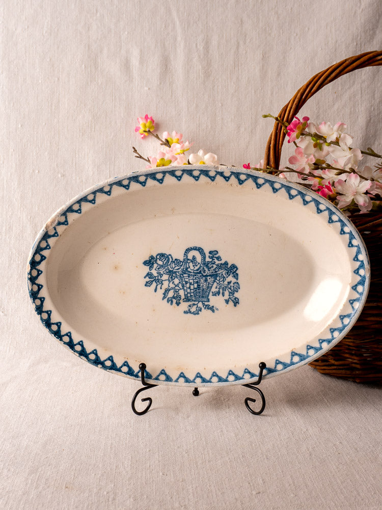 Antique French faience oval plate by Bertrand Opaque Surfine with blue floral basket, displayed before artificial flowers.