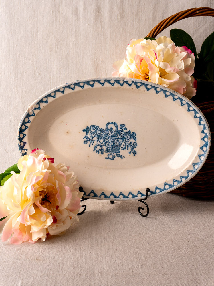 Antique French faience oval plate with blue flower basket on metal stand; pale flowers and wicker basket behind.