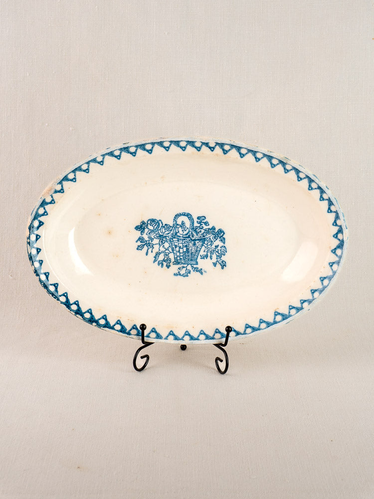 Antique French faience oval plate with blue zigzag border and central flower basket, on black stand, beige background.
