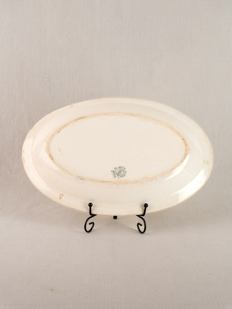 Antique French faience oval plate by Bertrand Opaque Surfine with blue floral basket, on black metal stand, light fabric background.