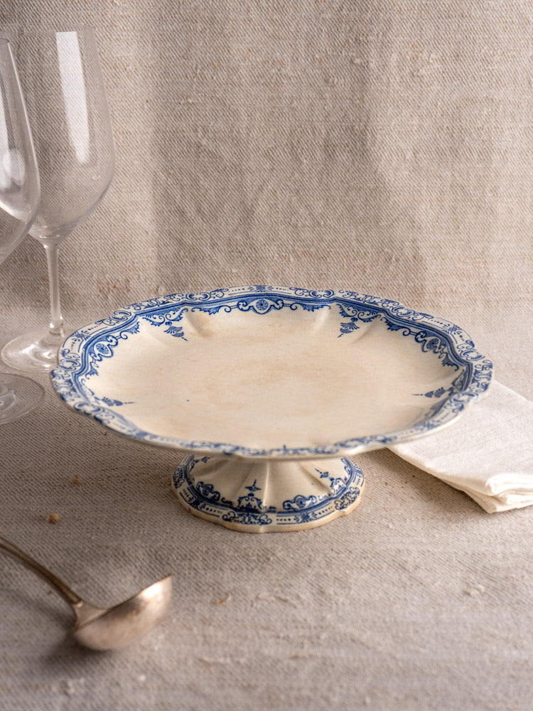 Antique French faience pedestal cake stand by E. Bourgeois Paris on textured beige cloth with wine glasses and silver spoon.