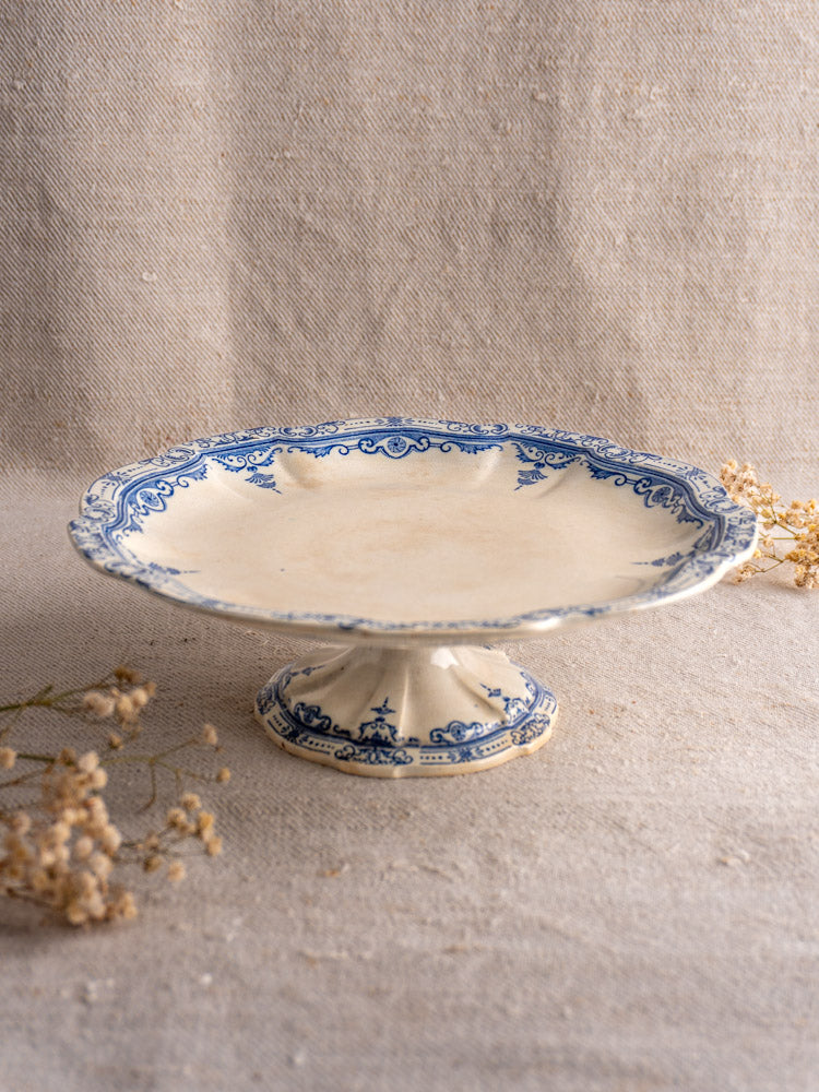 Antique French faience pedestal cake stand by E. Bourgeois, blue floral edge, on beige fabric with dried flowers.