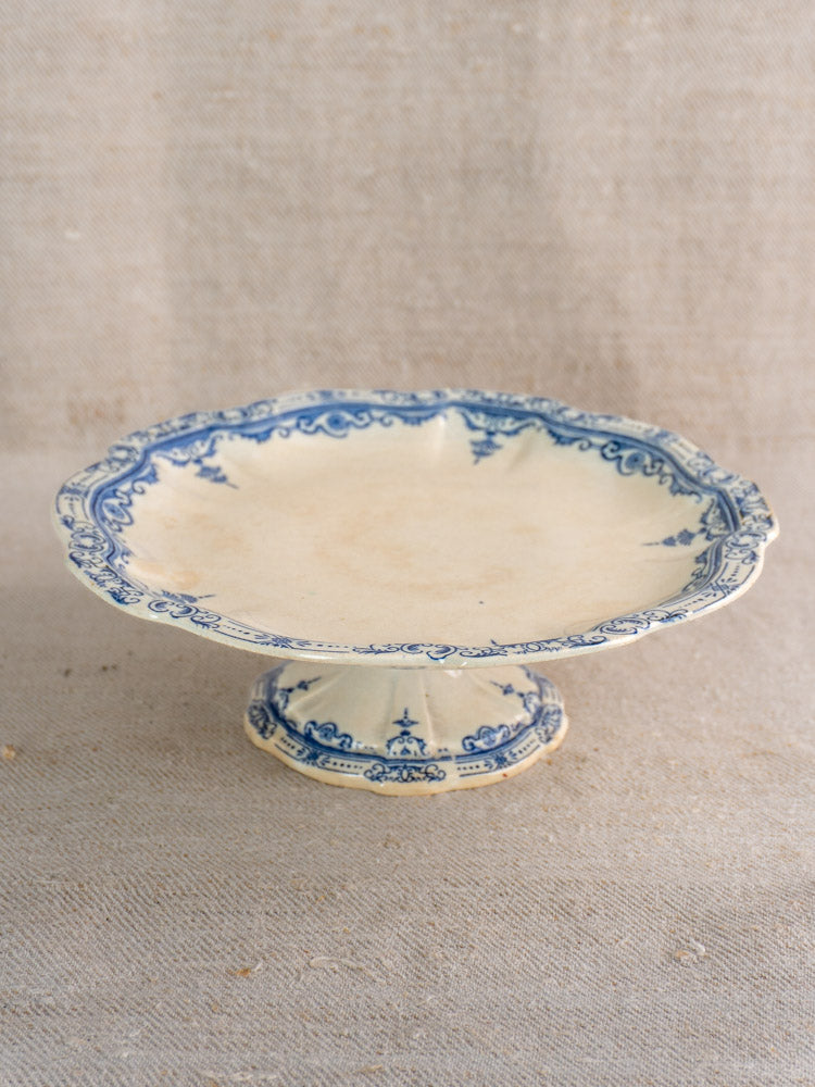 Antique French faience pedestal cake stand by E. Bourgeois Paris, scalloped edge, blue motifs on beige, on neutral fabric.