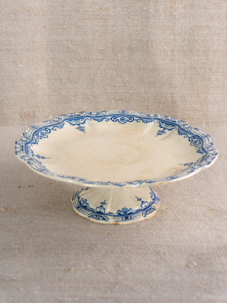 Antique French faience pedestal cake stand by E. Bourgeois with scalloped edges and blue floral scroll rim on beige fabric.