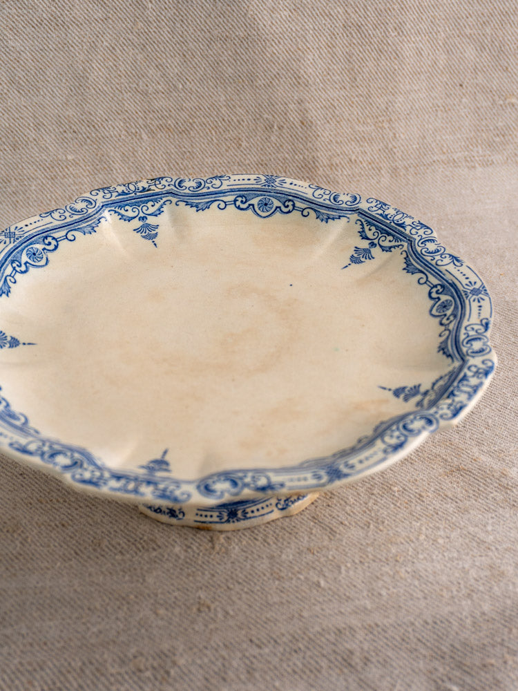 Antique French faience pedestal cake stand by E. Bourgeois Paris with scalloped edge, blue floral rim on neutral fabric background.