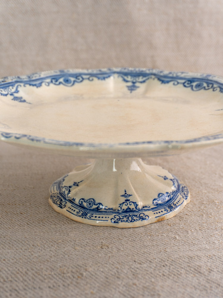 Antique French faience pedestal cake stand by E. Bourgeois, blue rim and base, on neutral textured fabric.