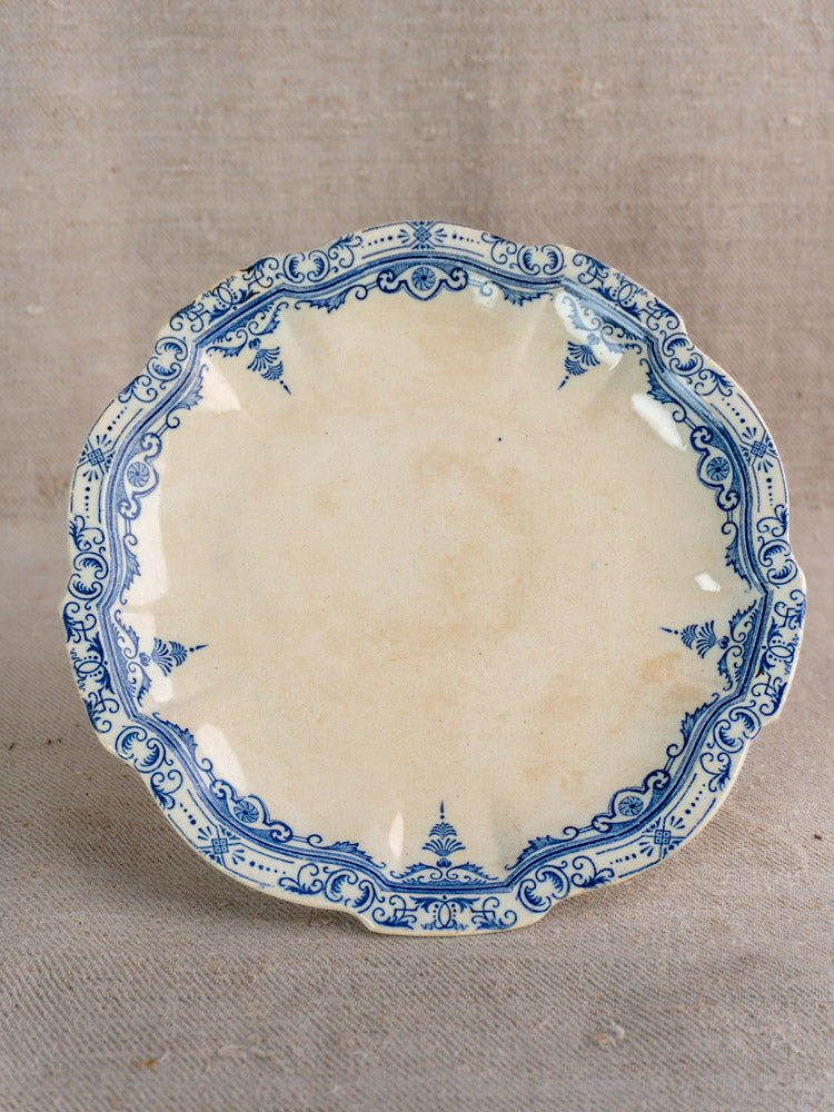 Antique French pedestal cake stand in faïence fine with blue floral scrolls, scalloped edges, on textured beige fabric background.