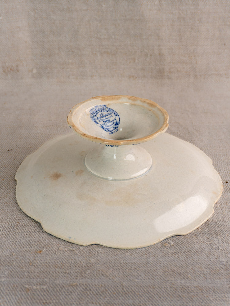 Antique French faience pedestal cake stand upside down, blue E. Bourgeois Paris Grand Dépôt mark visible on beige fabric.