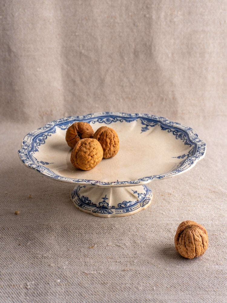 Antique French faience pedestal cake stand by E. Bourgeois Paris, scalloped edge, blue motifs on beige, on neutral fabric.
