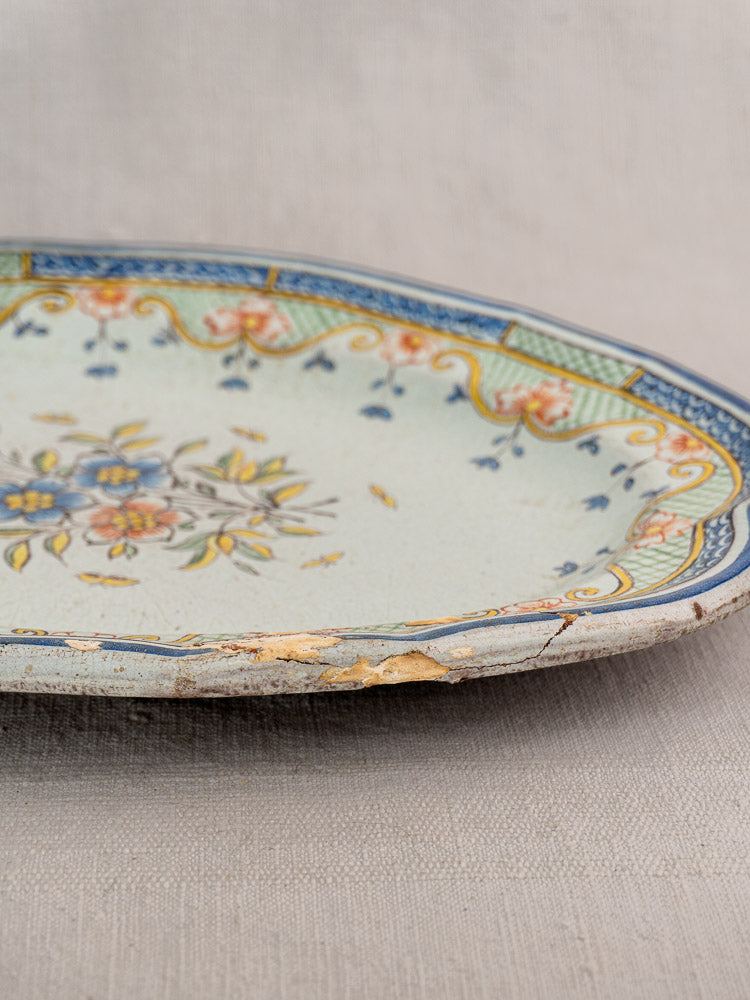 Antique French Orléans faience platter with floral polychrome design, colorful borders, chips, rim crack on fabric background.