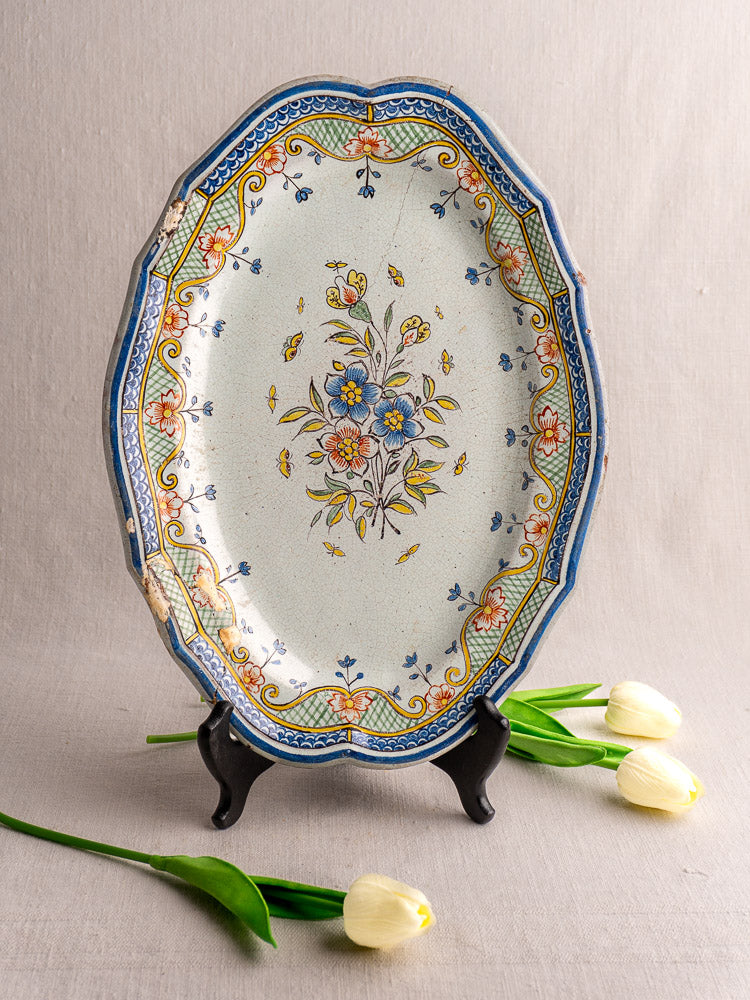 Antique French Orléans faience platter with polychrome floral motifs on black stand, white tulips on gray fabric nearby.