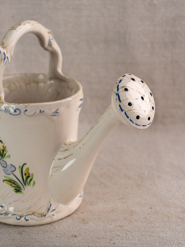 Vintage French faience watering can with hand-painted blue floral design on neutral fabric background.