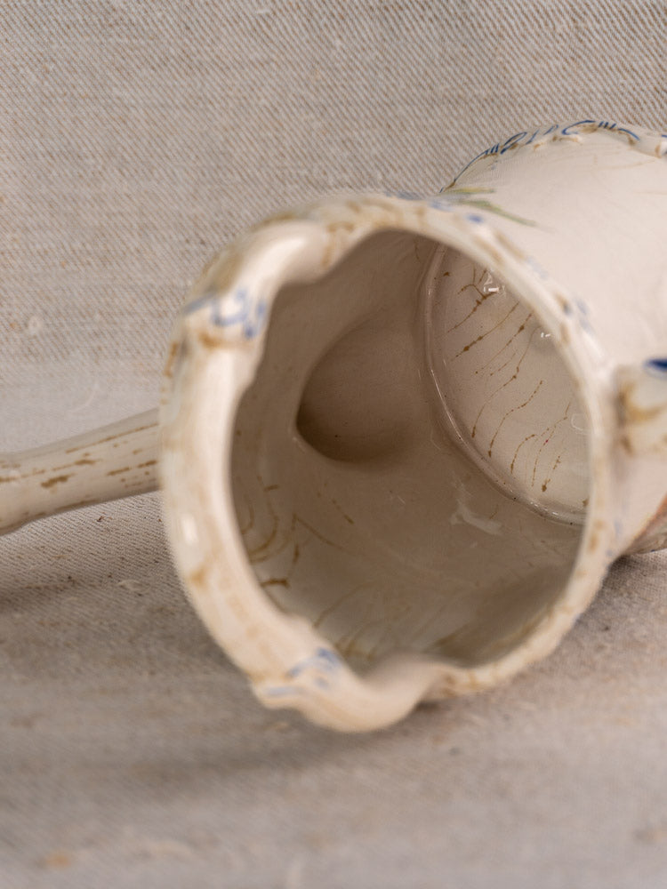 Vintage French faience watering can with scalloped rim and crackle lines on beige fabric background.