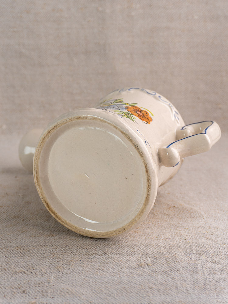French faience watering can with hand-painted floral design, shown on textured beige fabric.