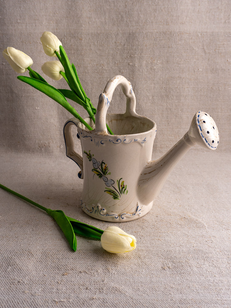 Vintage French faience watering can with hand-painted florals, holding three white tulips on beige surface.