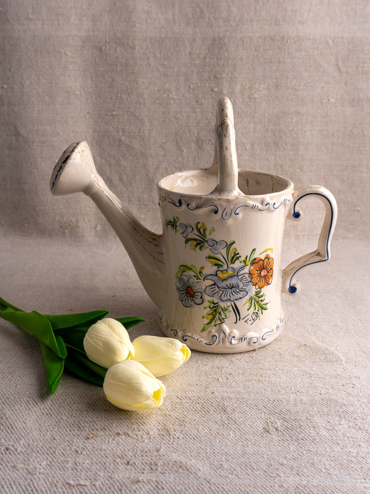 Vintage French faience watering can with hand-painted floral motifs beside yellow tulips on beige fabric.