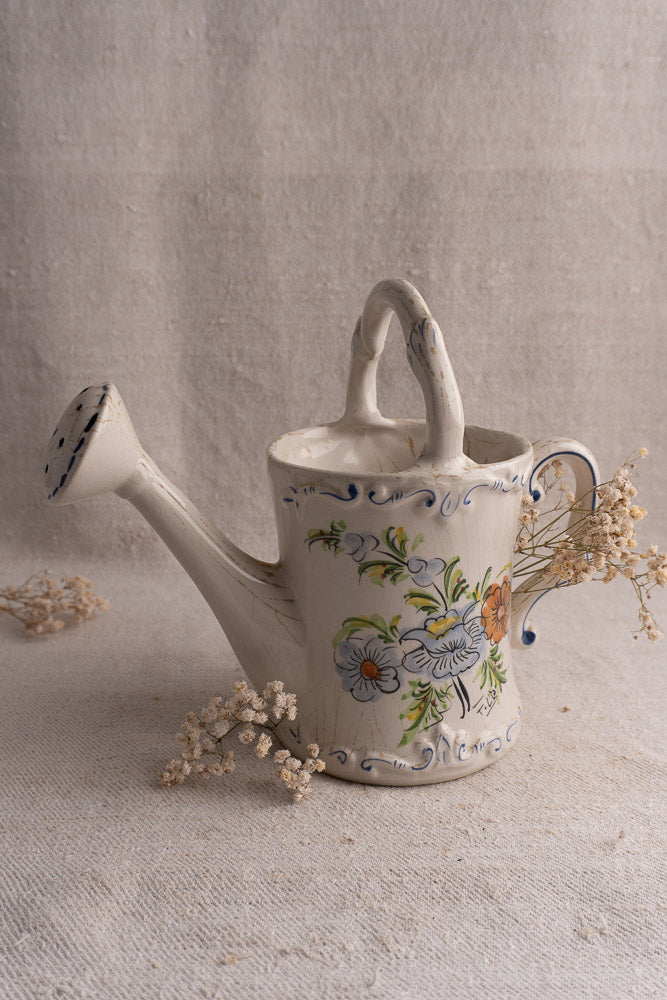 Hand-painted vintage French faience watering can on textured cloth, surrounded by dried white flowers.