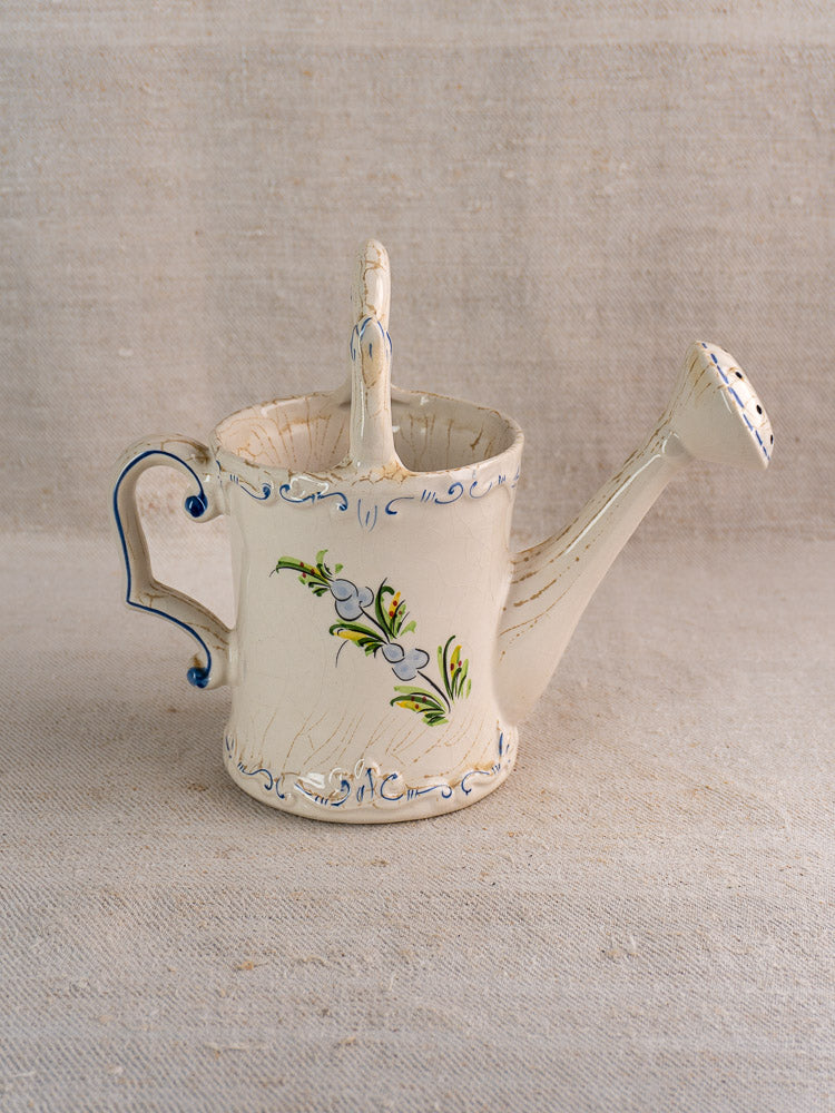 Vintage French faience watering can, cream with blue and green hand-painted florals, on beige textured surface.