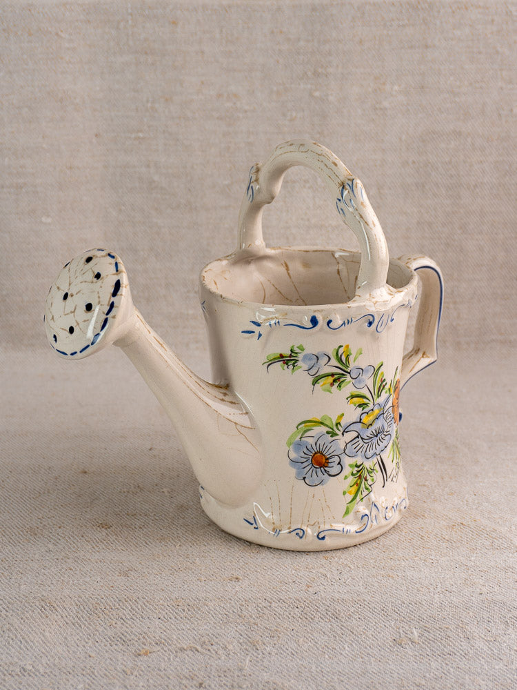 Vintage French faience watering can with blue and yellow floral motifs on beige cloth background.