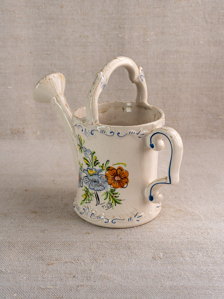 Vintage French faience watering can with hand-painted blue and orange flowers on beige fabric.