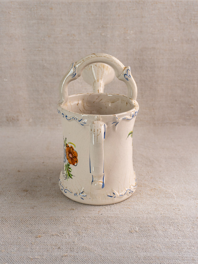Vintage French faience watering can with hand-painted floral design on a neutral fabric background.