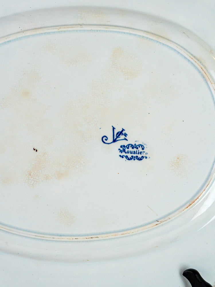 Underside of white ceramic plate with blue “Moustiers” mark, decorative details, and light age spots.