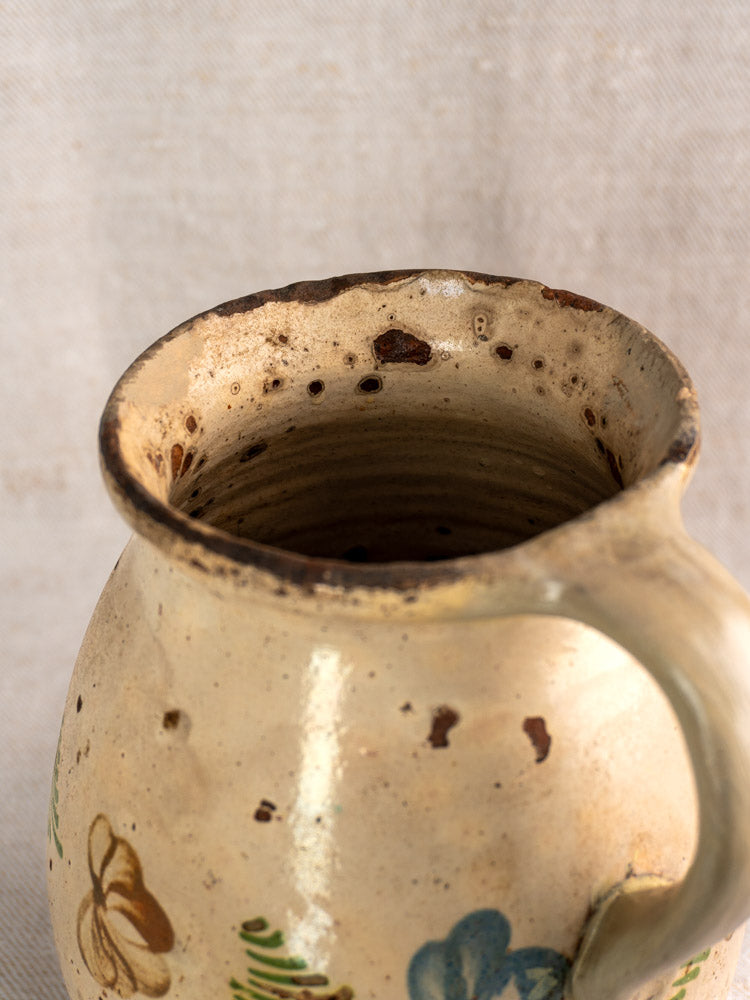 Antique French faience pitcher, mid-19th century, hand-painted polychrome florals, chipped rim on textured beige fabric.