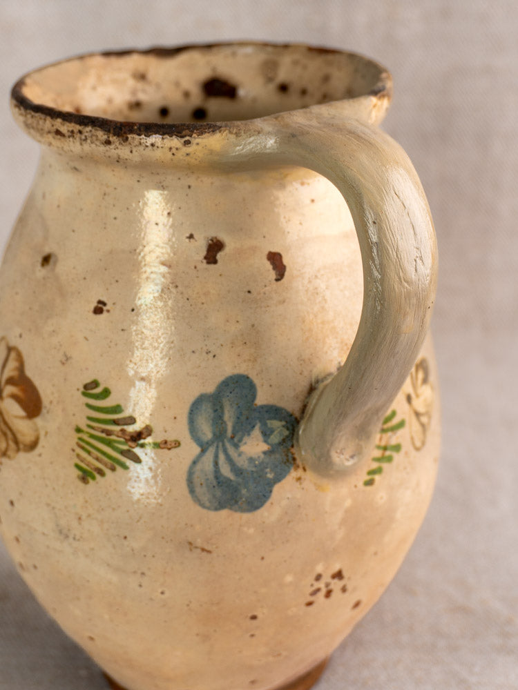 Antique French faience pitcher with curved handle, hand-painted floral motifs, and aged speckled glaze on fabric background.