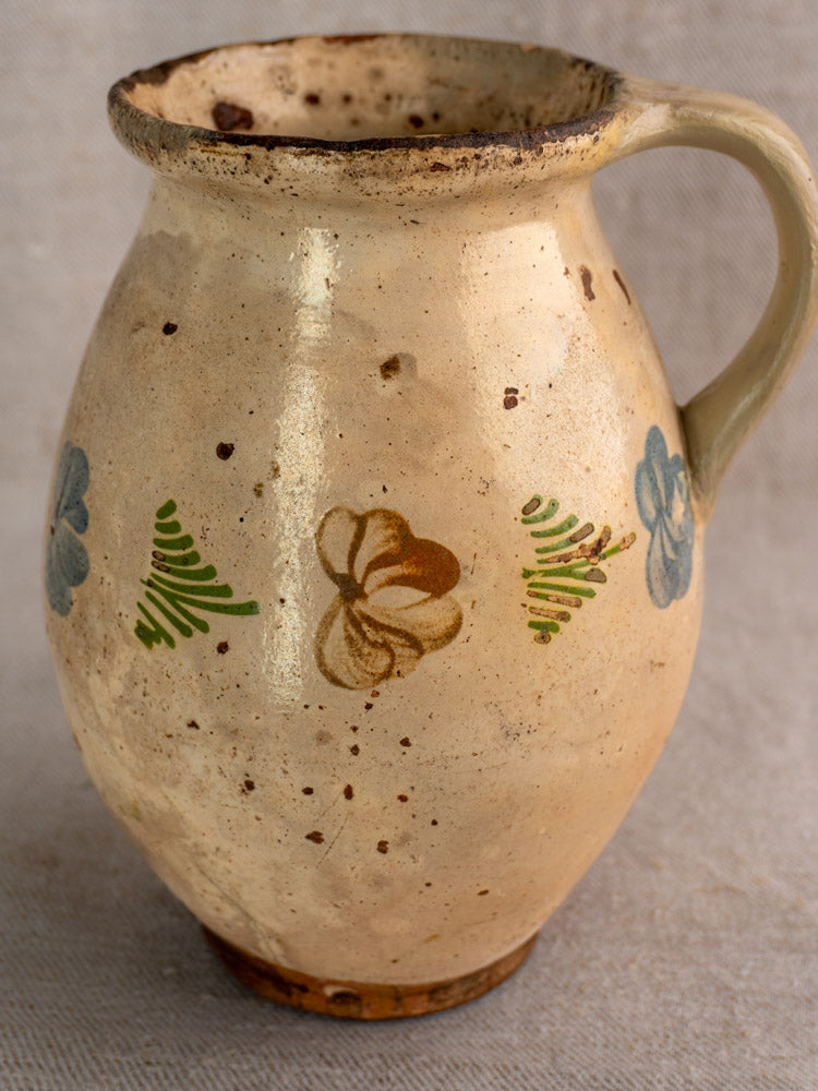 Antique French faience pitcher with hand-painted polychrome floral motifs and a handle on a textured light gray surface.