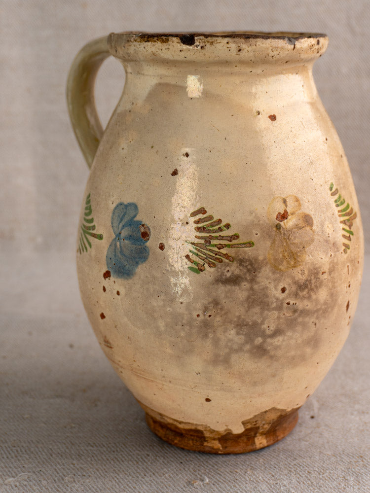 Antique French faience pitcher with handle, hand-painted green leaves and blue-yellow florals on creamy body, on beige surface.
