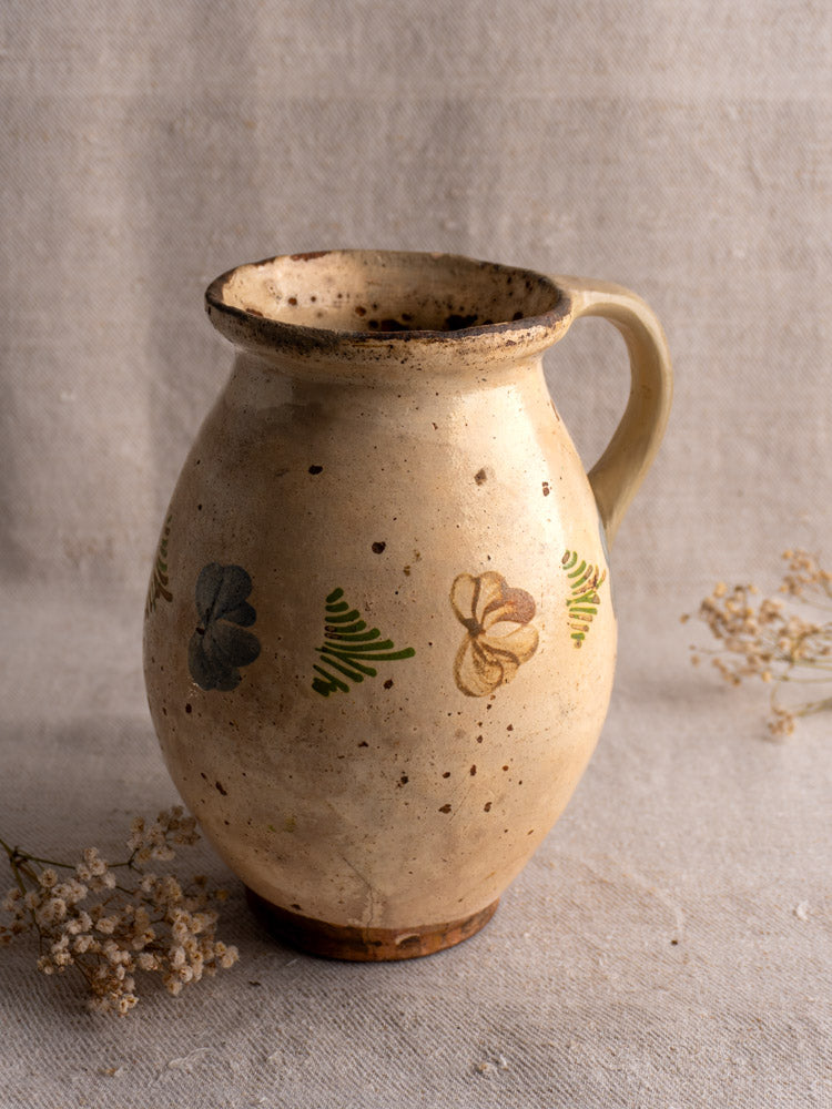 Antique French faience pitcher with polychrome floral motifs and single handle, cream with rustic wear on neutral fabric.