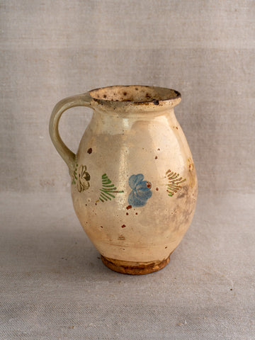Antique French faience pitcher with polychrome floral motifs and single handle, cream with rustic wear on neutral fabric.
