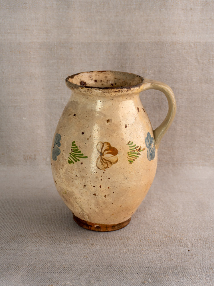 Antique French faience pitcher with hand-painted polychrome floral and brown motifs, rustic beige body, on neutral fabric background.