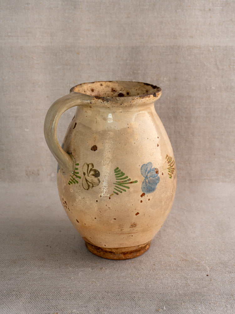 Antique French faience pitcher, mid-19th century, beige with hand-painted green, blue, and brown floral motifs on fabric background.
