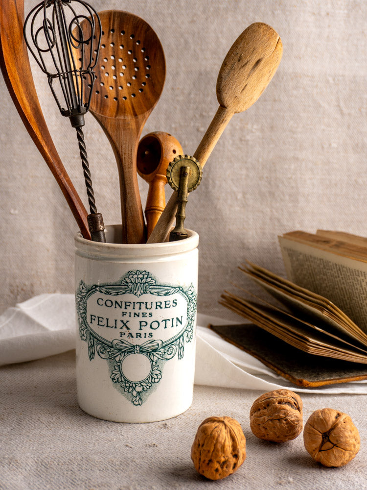 Antique French Felix Potin jam pot marked "Confitures Fines Paris" with vintage utensils, walnuts, and open book on linen.