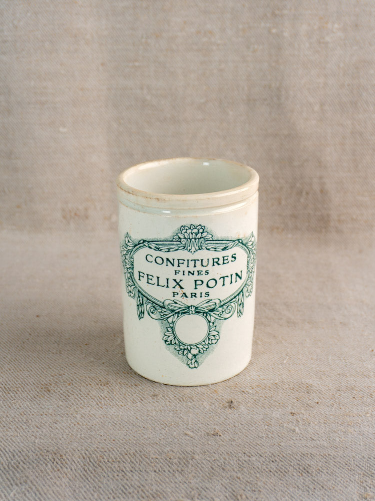 Antique French Felix Potin faience jam pot with blue "Confitures Fines Paris" label on textured beige fabric.