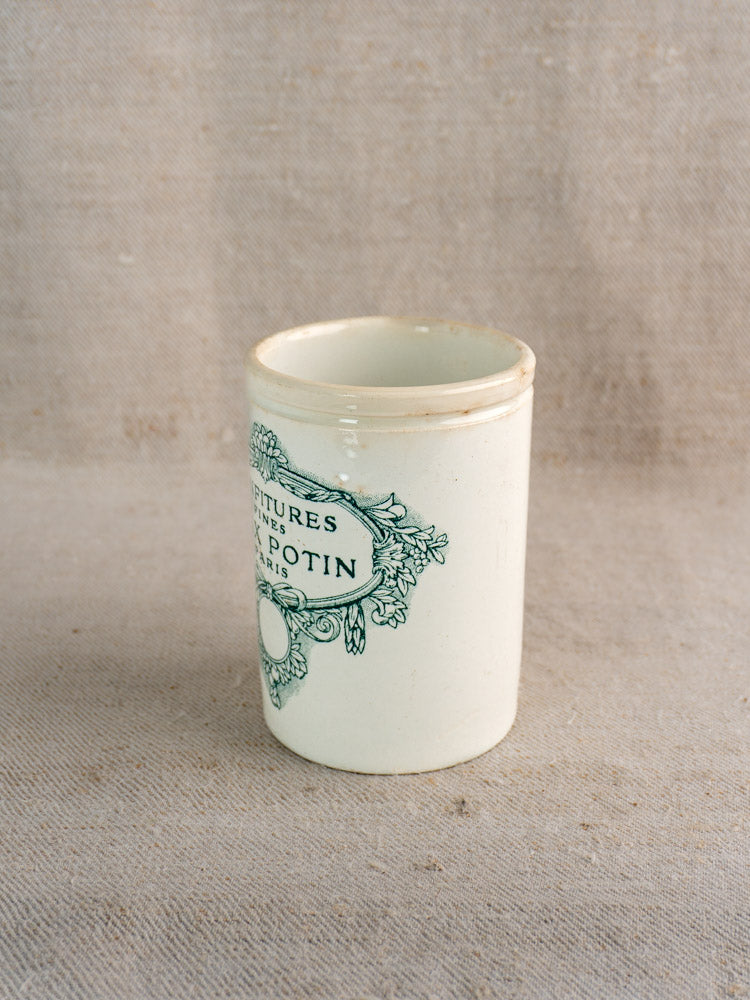 Antique French Felix Potin confiture pot with green label on white ceramic, set on textured beige fabric.