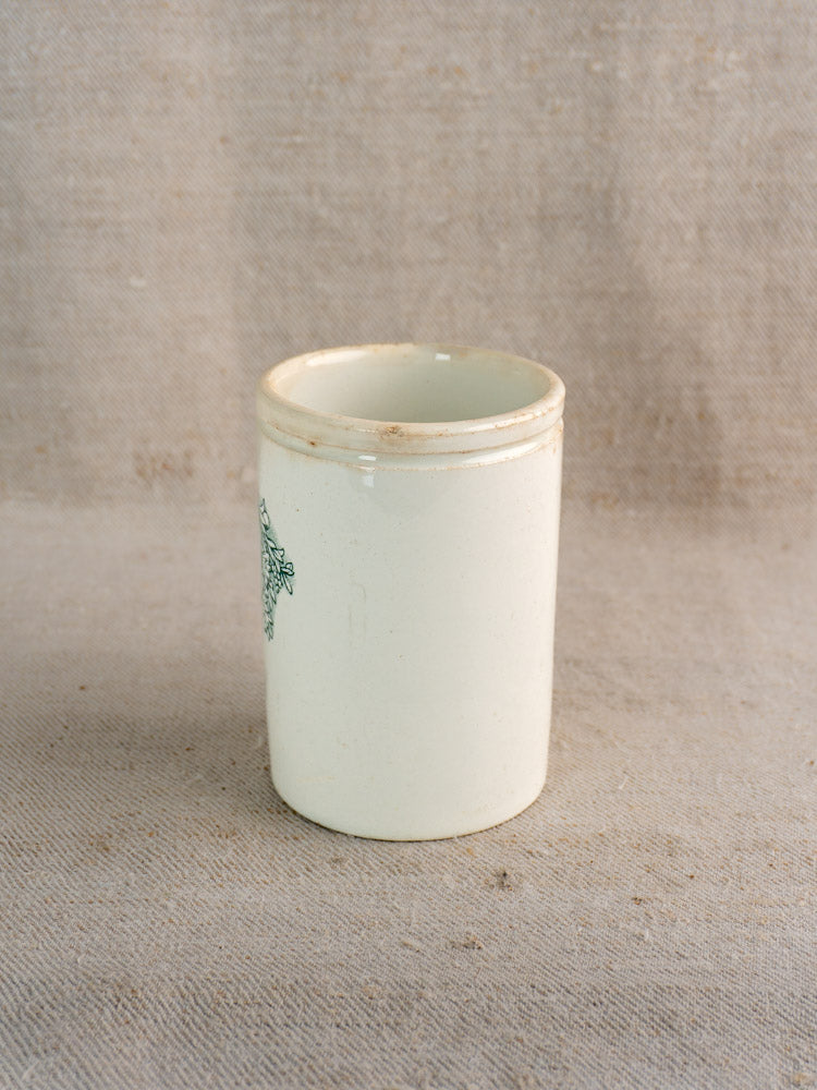 Antique French Felix Potin jam pot in white faience with worn rim and green design, on beige fabric.