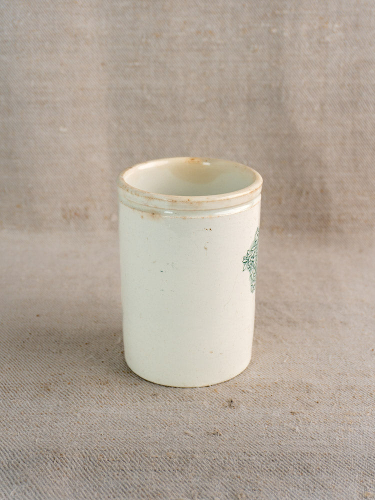 Antique French Felix Potin ceramic jam pot, off-white with green mark, cylindrical, gently worn rim on beige textured fabric.