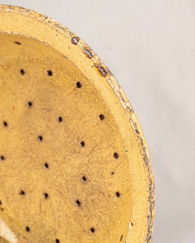 Antique French earthenware égouttoir with yellowish glaze, evenly spaced holes, and weathered rim on neutral background.