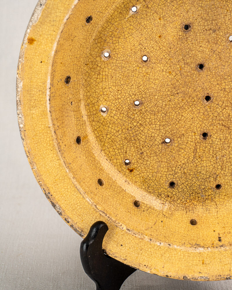 Antique French glazed earthenware fruit strainer, 19th century, yellow with cracks and holes, on black stand.