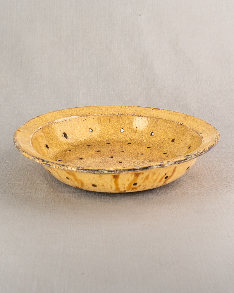 Shallow round yellow glazed earthenware égouttoir with textured surface and holes, on light-grey background.