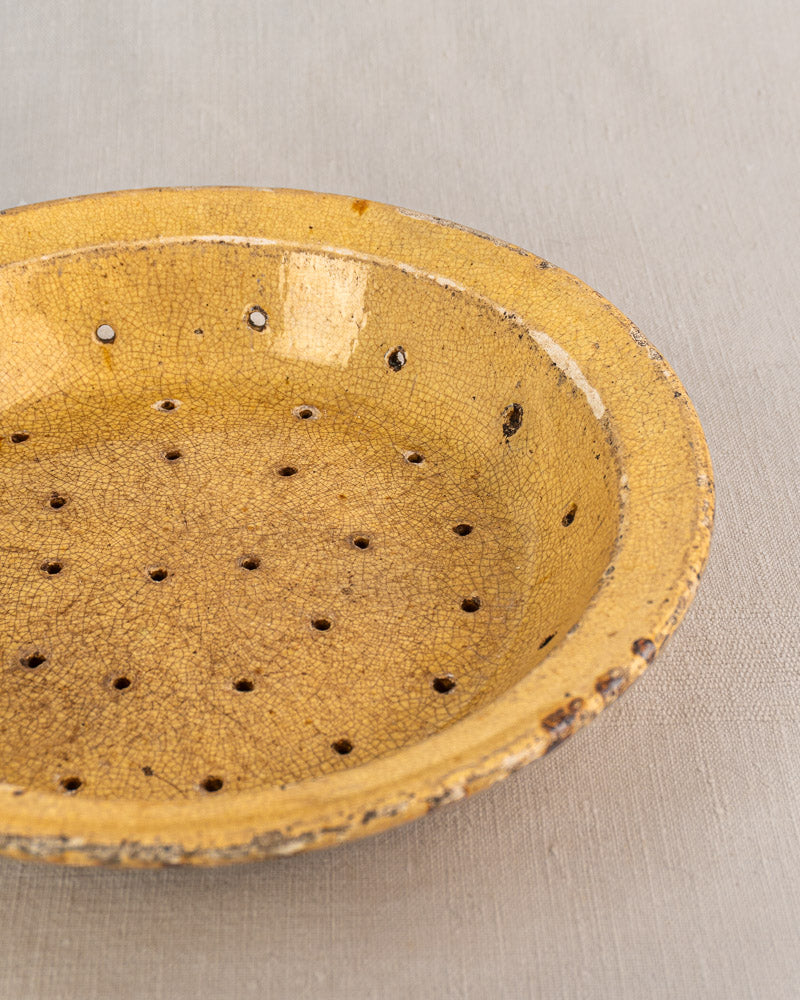 Antique French yellow glazed earthenware fruit strainer with crackled finish and holes on light gray fabric background.